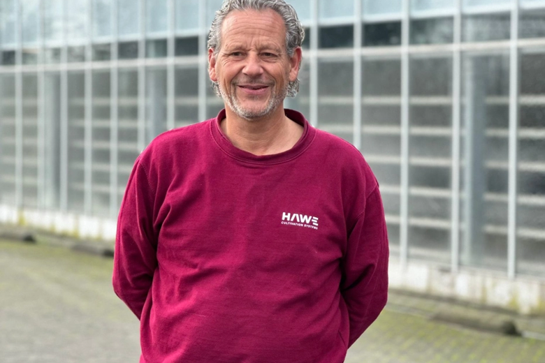 Mann in bordeauxfarbenem Hawe-Pullover vor einem Gewächshaus